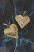 Two gold heart drop earrings with a textured finish resting on dark green succulent leaves.