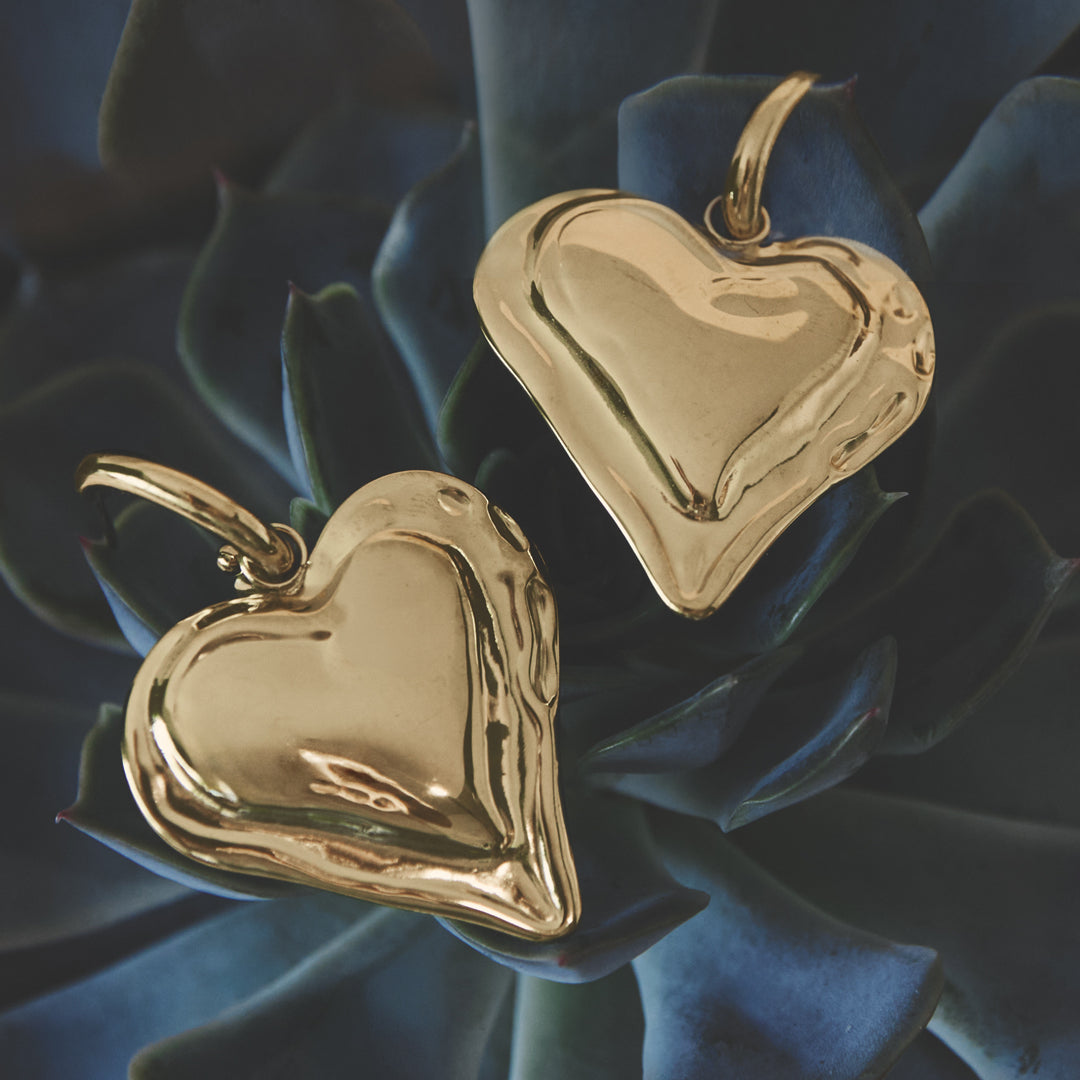 Two gold heart drop earrings with a textured finish resting on dark green succulent leaves.