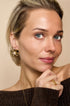 Gold flower stud earrings with pearl accents, worn by a woman with blue eyes and blonde hair.
