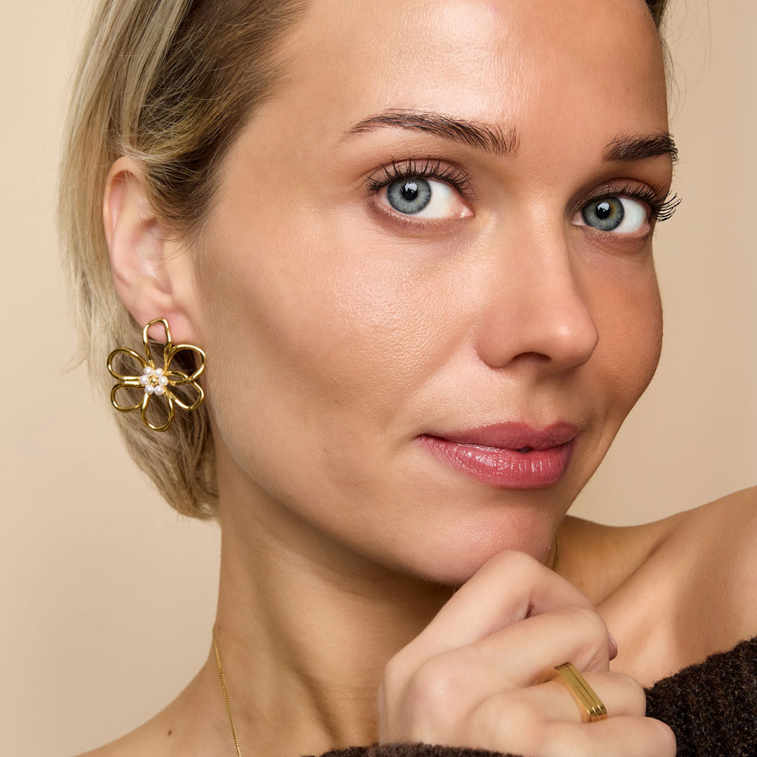 Gold flower stud earrings with pearl accents, worn by a woman with blue eyes and blonde hair.