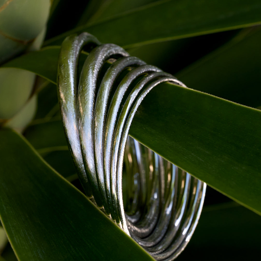 Silver Elara Bangle Bracelet with a brushed finish, 3 mm wide, resting on green leaves.