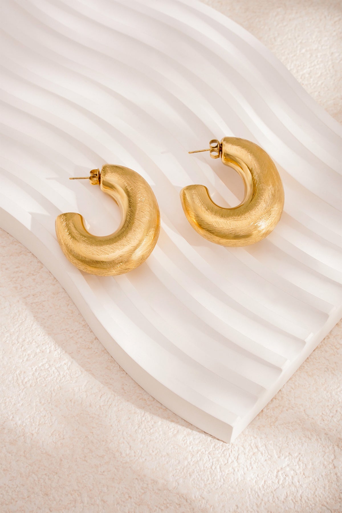 Gold Elysian Hoop Earrings with a brushed finish, displayed on a white wavy surface.