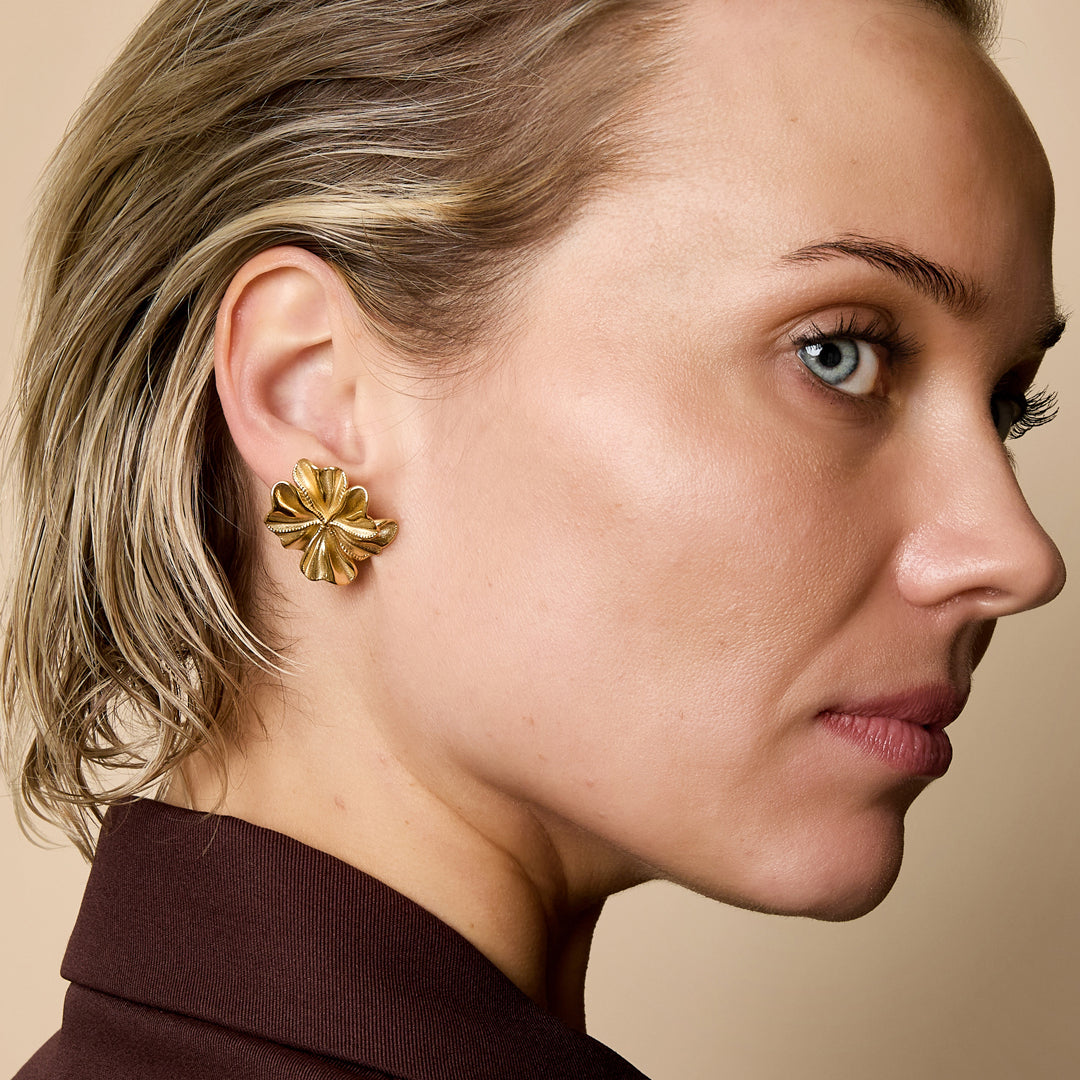 Gold-colored petal stud earrings with a textured surface, worn by a woman.