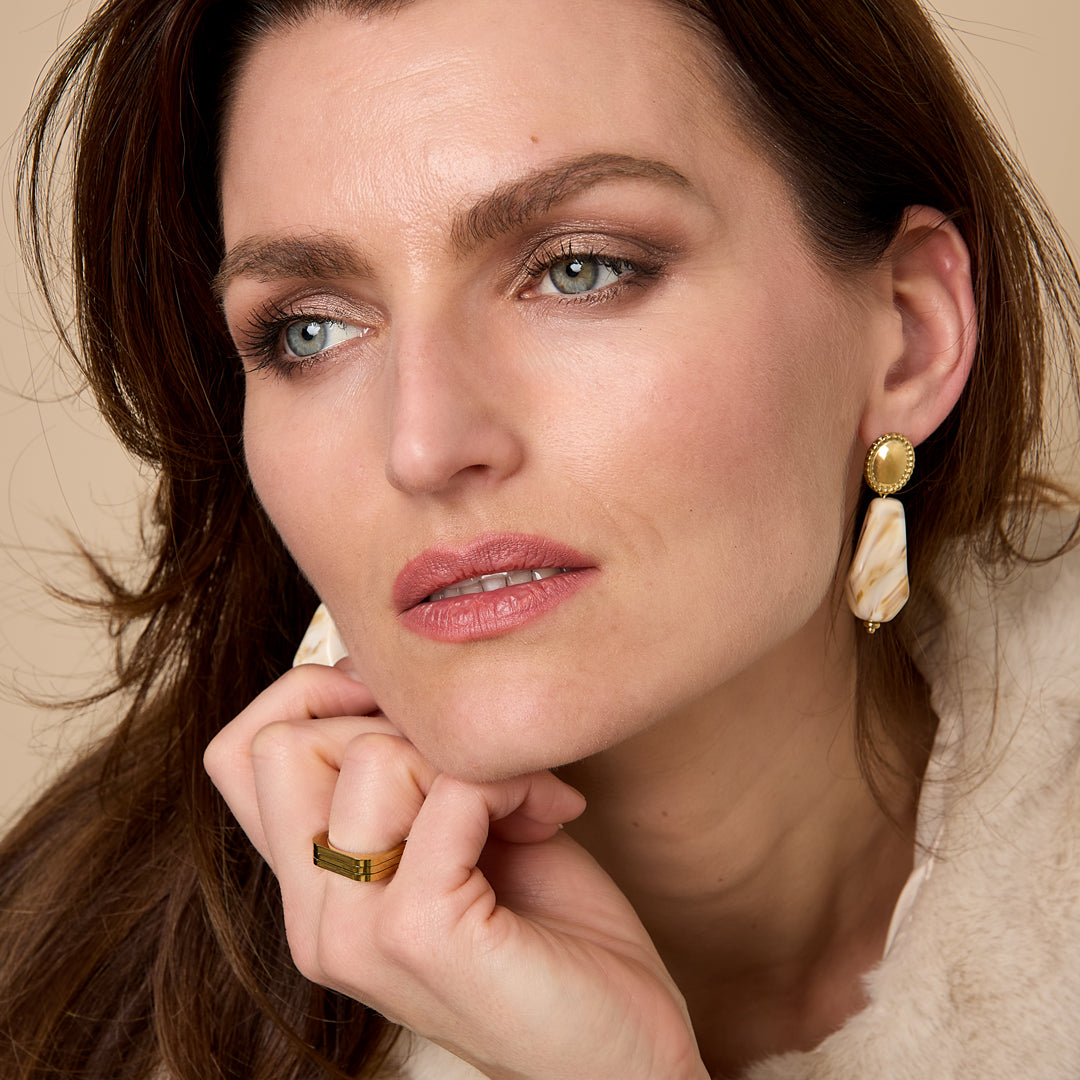 Woman wearing amber irregular earrings and a gold ring, with a beige fur coat.