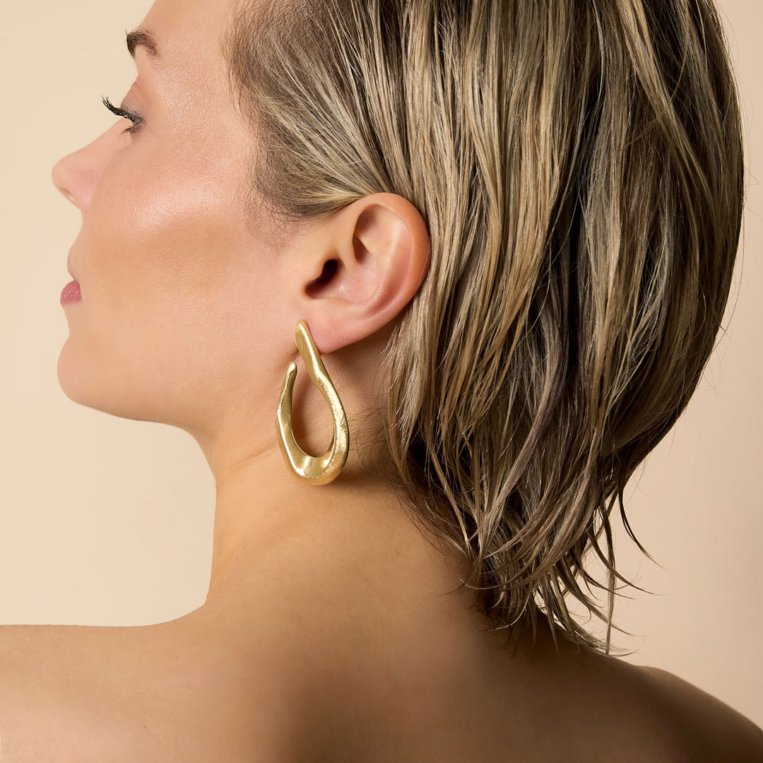 Gold-toned, irregular shaped hoop earrings on a woman's ear.