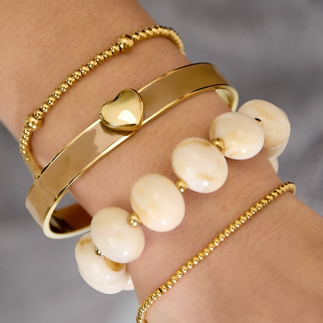 Gold bangle with a heart charm, layered with beaded bracelets and cream-colored beads.