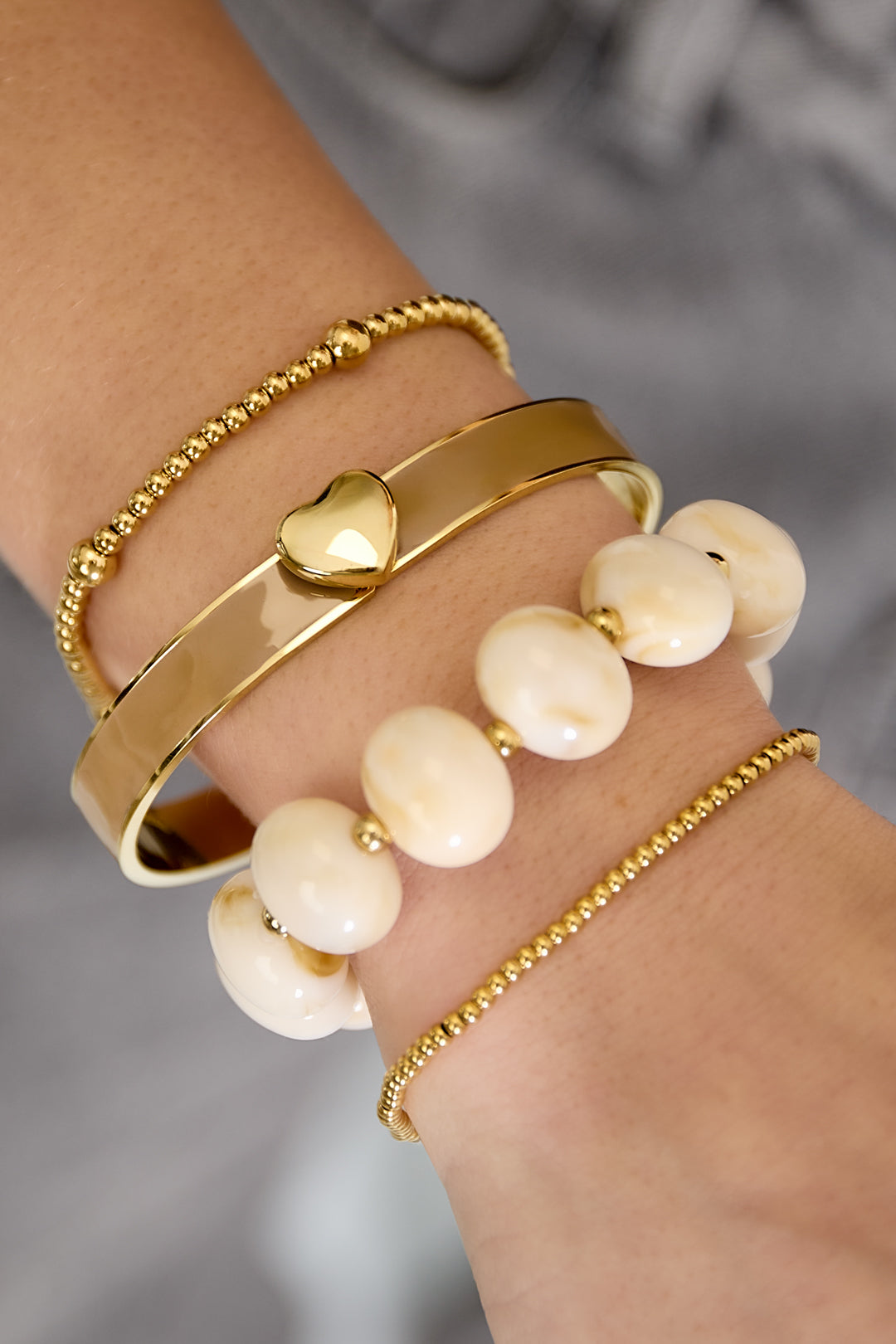 Gold bangle with a heart charm, layered with beaded bracelets and cream-colored beads.
