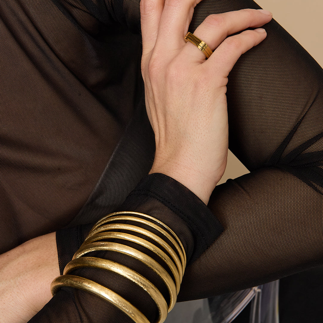 A stack of gold Elara bangle bracelets, 10 mm wide, worn on a wrist with a gold ring.