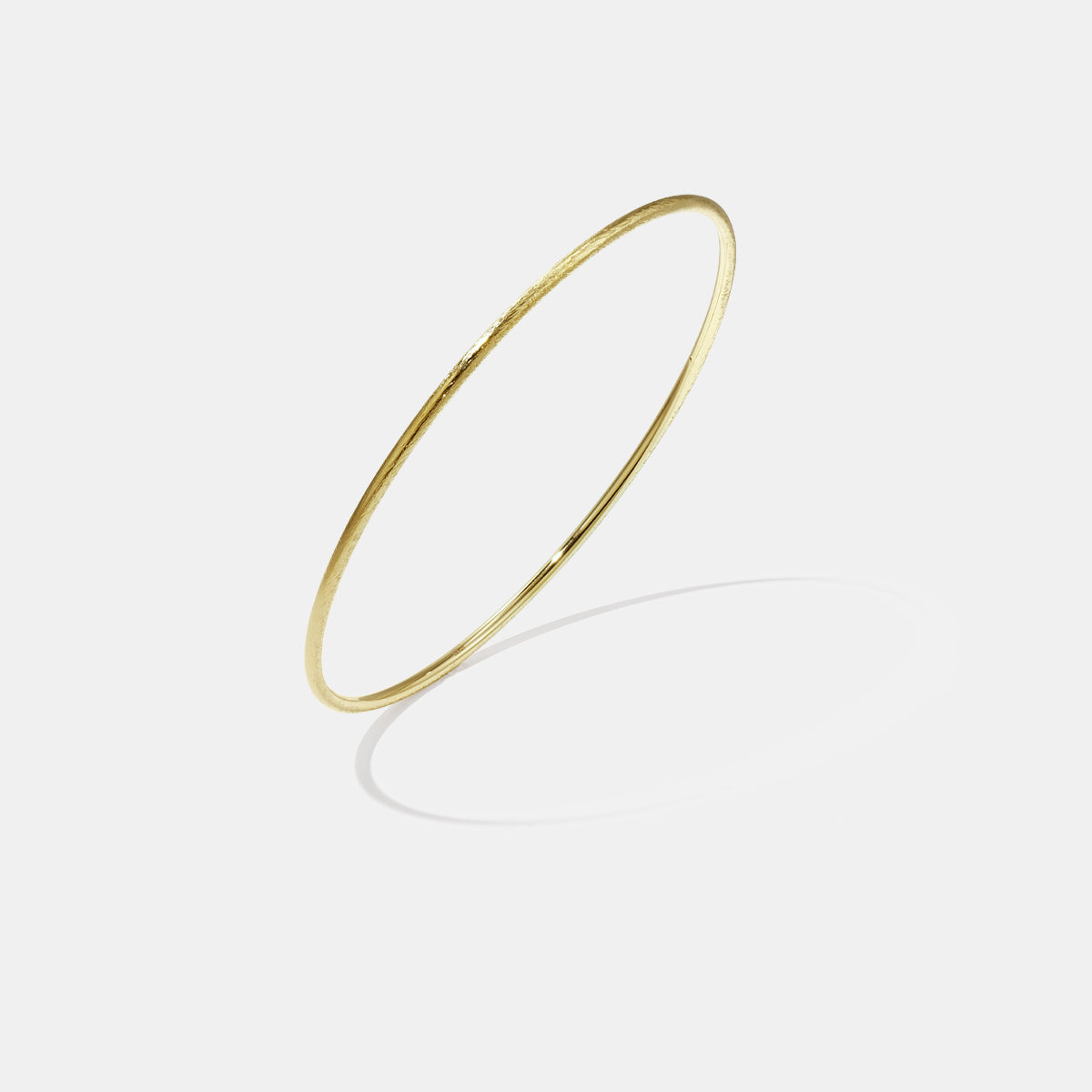 Thin gold bangle bracelet with a brushed finish, casting a shadow on a light grey background.