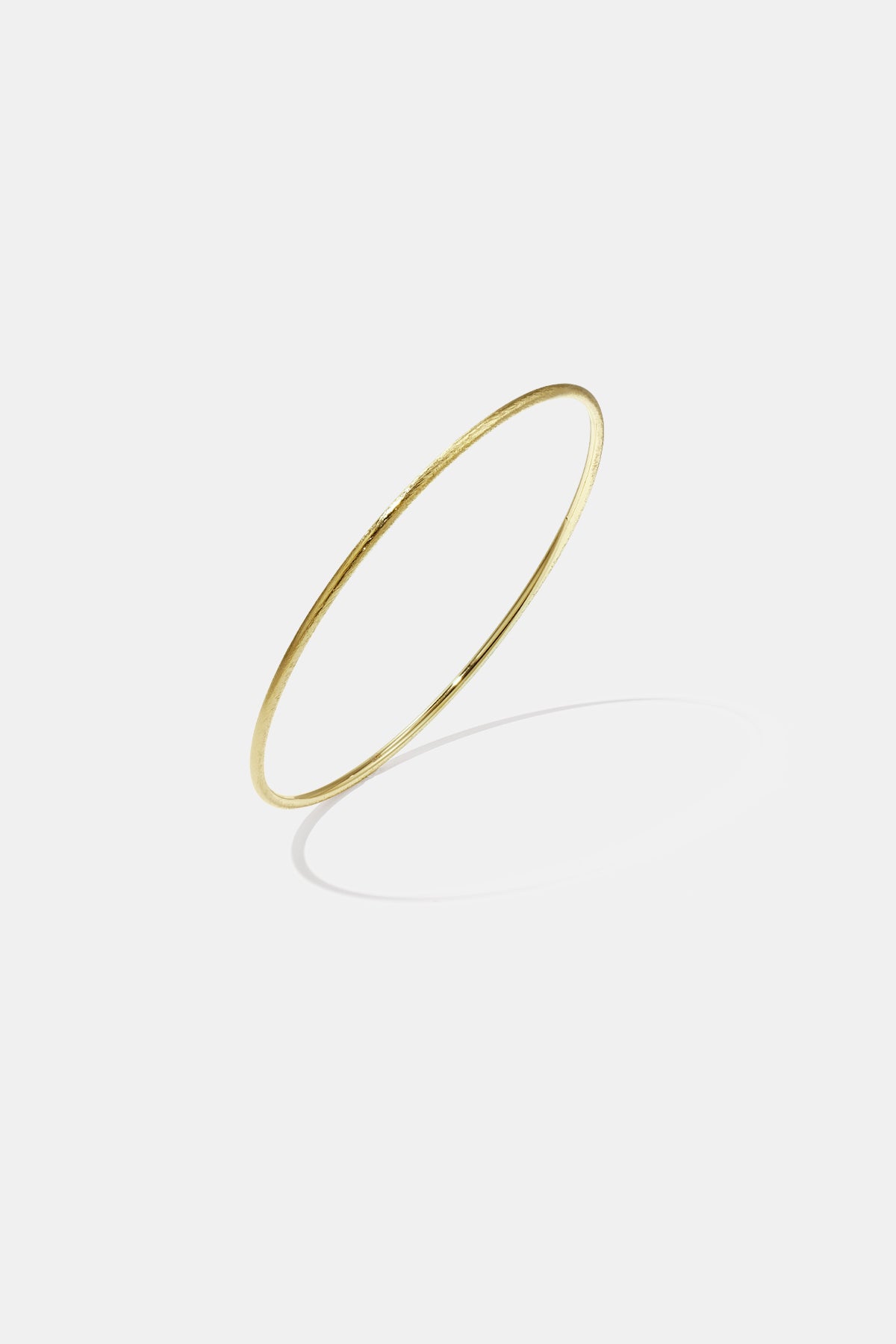 Thin gold bangle bracelet with a brushed finish, casting a shadow on a light grey background.