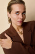 Gold heart drop earrings on a woman wearing a brown blazer and pearl necklace.