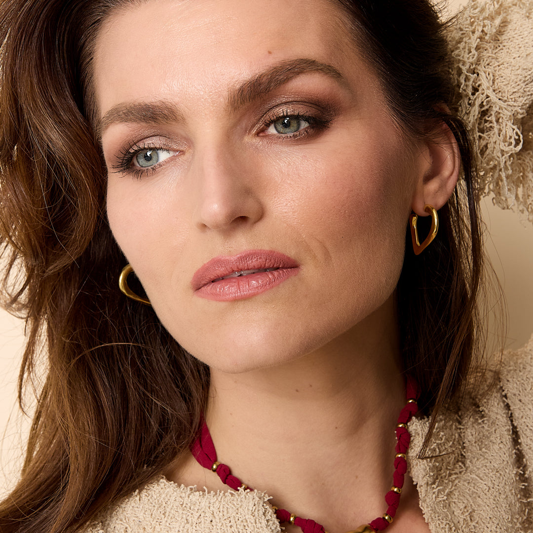 Gold heart hoop earrings on a woman wearing a beige textured jacket and a red necklace.