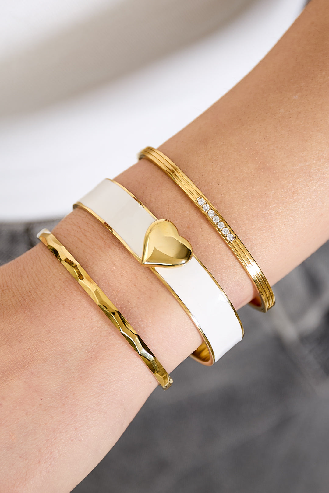 Three gold bangles on a wrist: one with a white band and gold heart, one with small diamonds, and one textured.