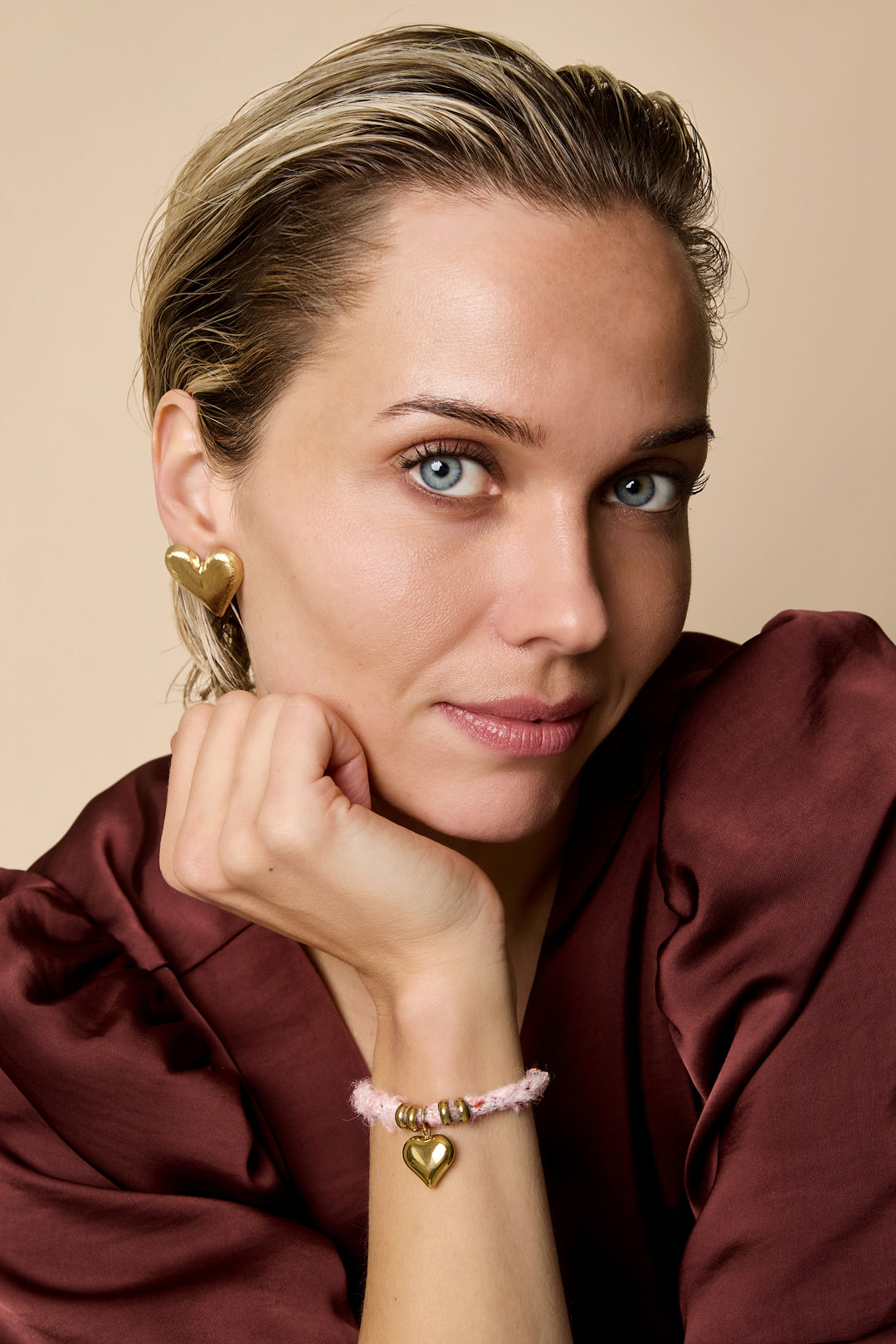 A woman wears a coffee brown/pink rope bracelet with a gold heart charm and gold rings.