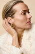 Gold-toned irregular hoop stud earrings on a woman's ear, styled with a textured white jacket.