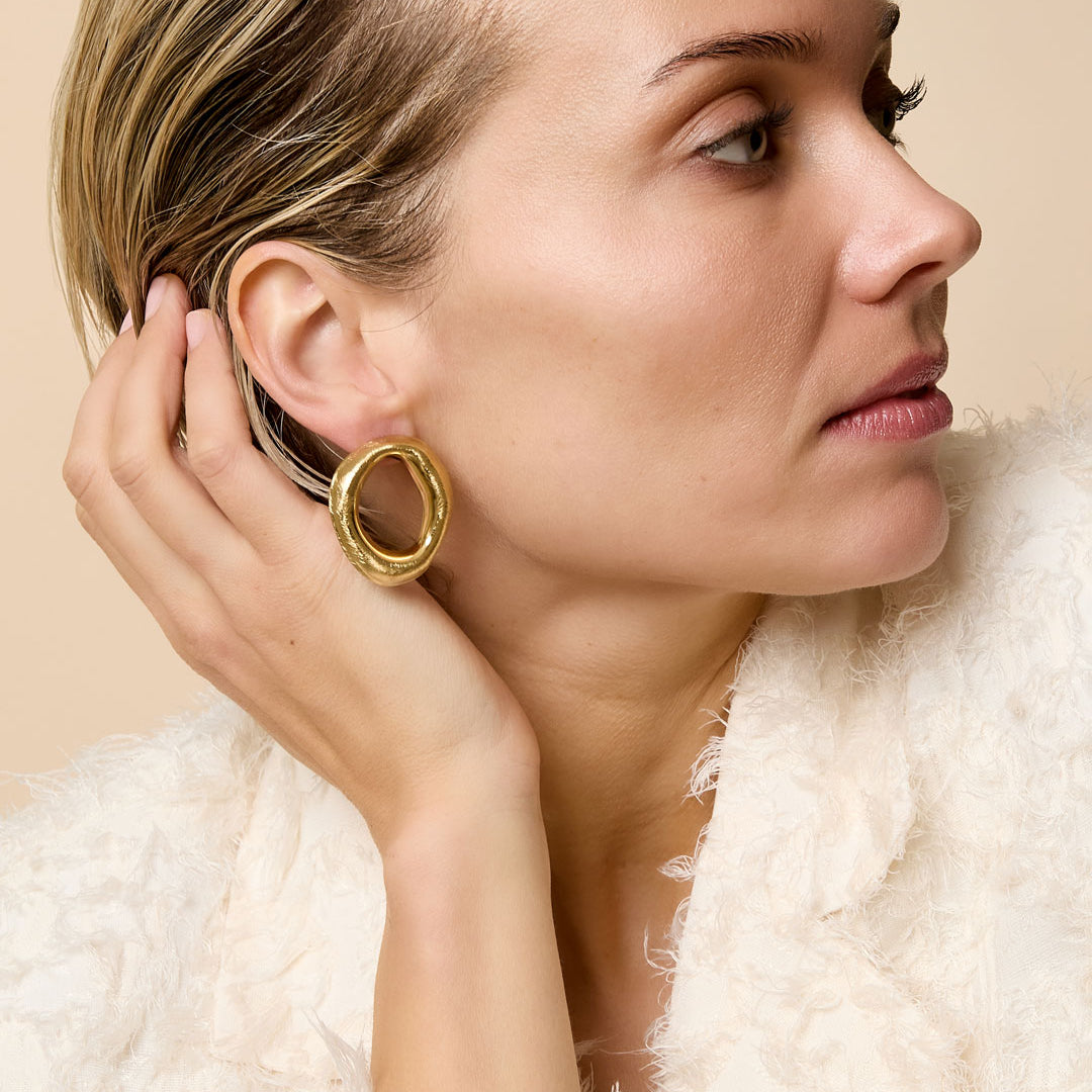 Gold-toned irregular hoop stud earrings on a woman's ear, styled with a textured white jacket.