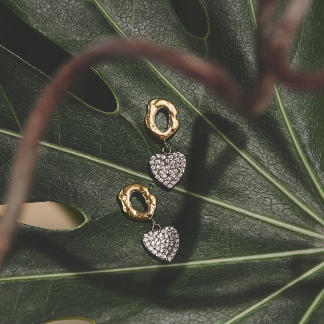 Gold drop earrings with textured rings and pave zircon heart charms on a green leaf.
