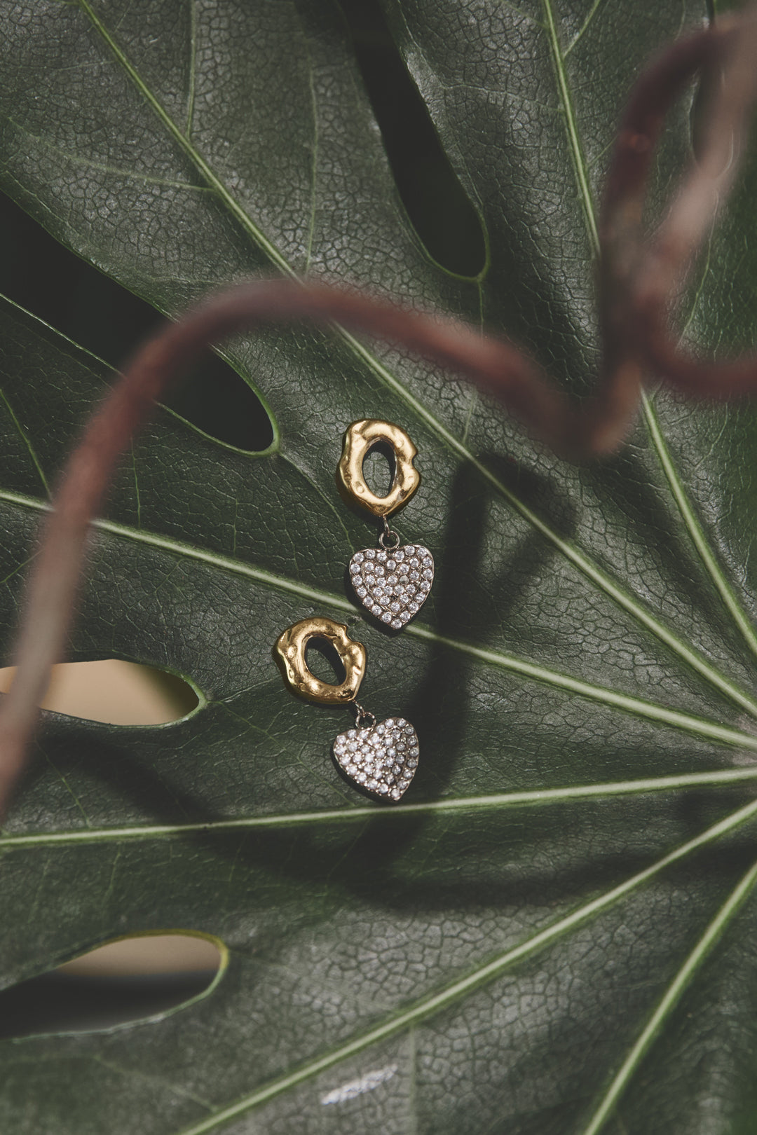 Gold drop earrings with textured rings and pave zircon heart charms on a green leaf.