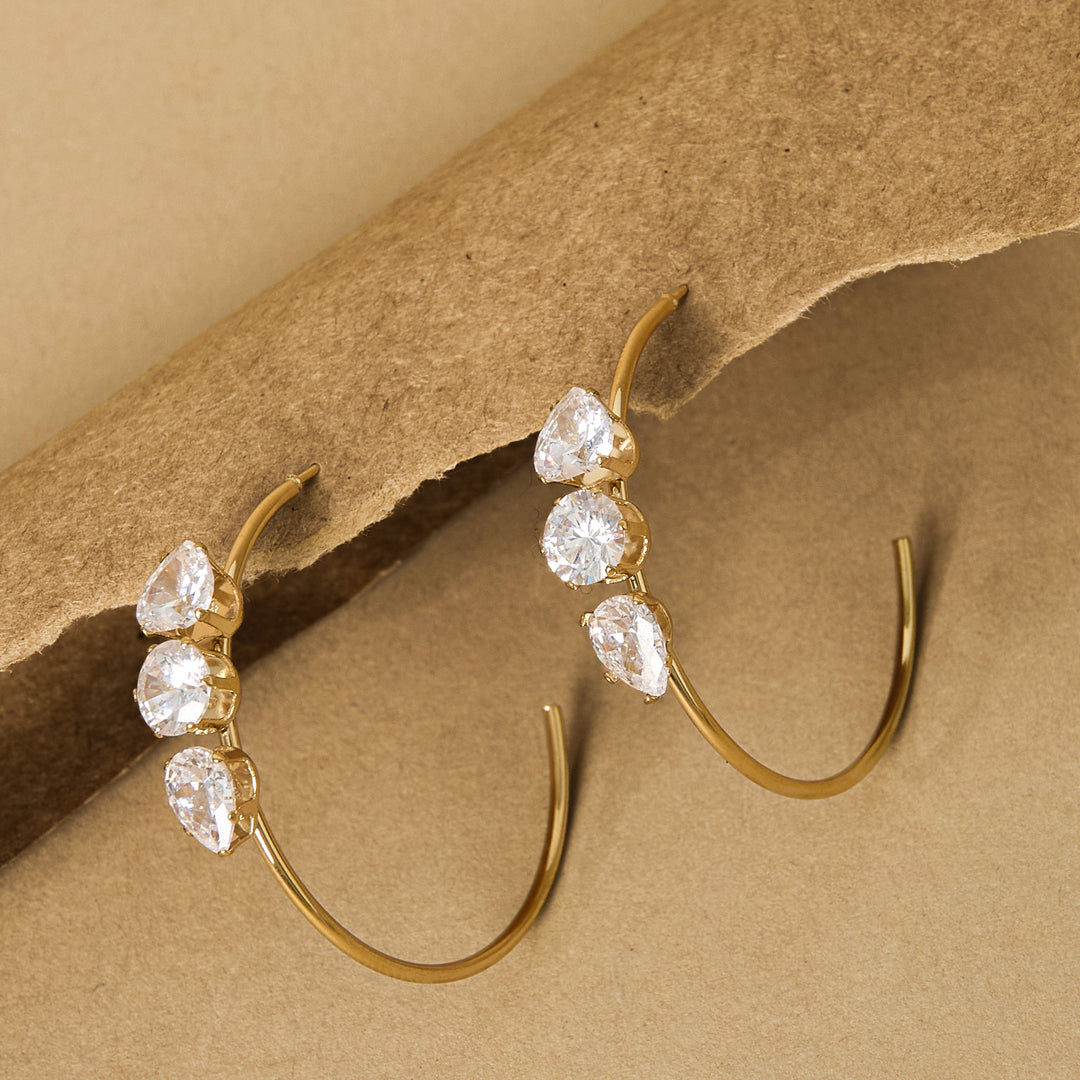 Gold hoop earrings with three clear stones each, set against a brown background.