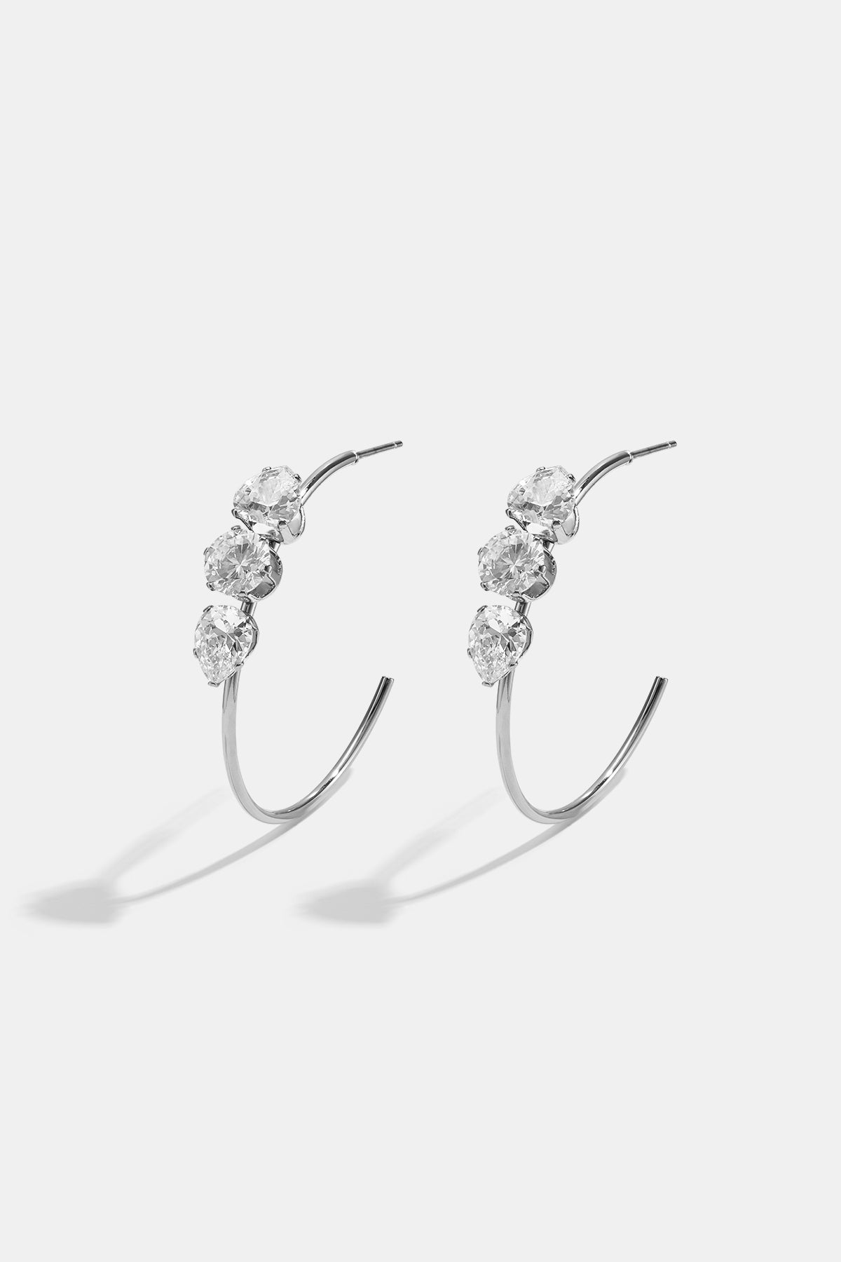 Silver hoop earrings with three clear stones each, casting shadows on a white background.