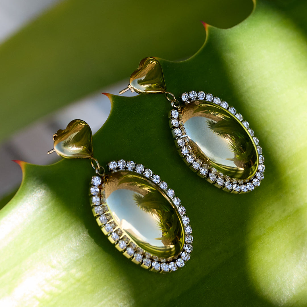 Gold oval statement earrings with rhinestones and heart-shaped tops on a green leaf.
