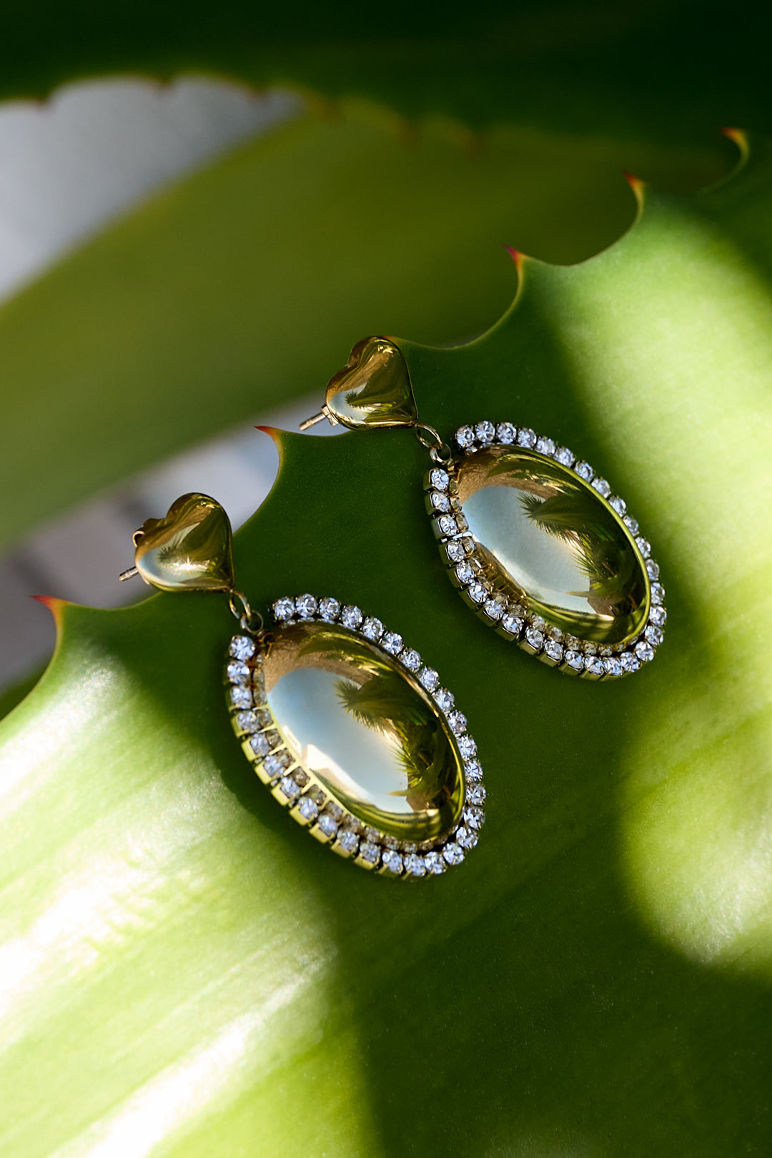 Gold oval statement earrings with rhinestones and heart-shaped tops on a green leaf.